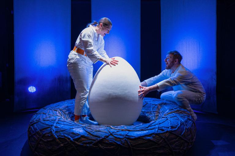 Två skådespelare står i ett stort bo och håller i ett stort vitt ägg i blått scenljus.