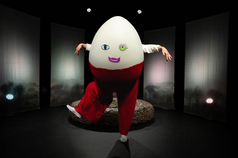 Person i äggkostym med ansikte och röda byxor står framför boet på scenen.