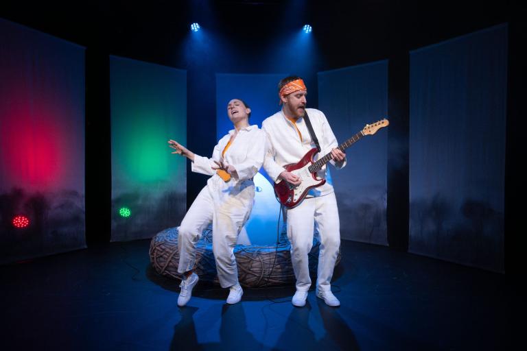 Två skådespelare showar på scen; en sjunger och en spelar röd elgitarr. Rött och grönt ljus.