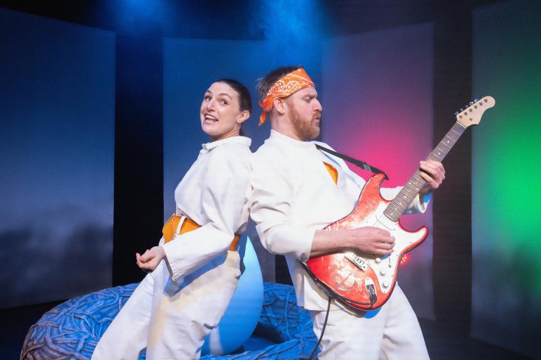 Två skådespelare rygg mot rygg framför ägget, den ena spelar elgitarr i färgstarkt scenljus.