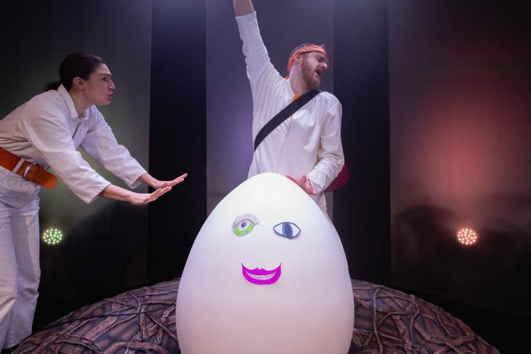 Skådespelare på scen med stort vitt ägg i ett bo – en person sträcker upp ena armen med inlevelse medan en annan står bredvid och ser ut att hyscha..