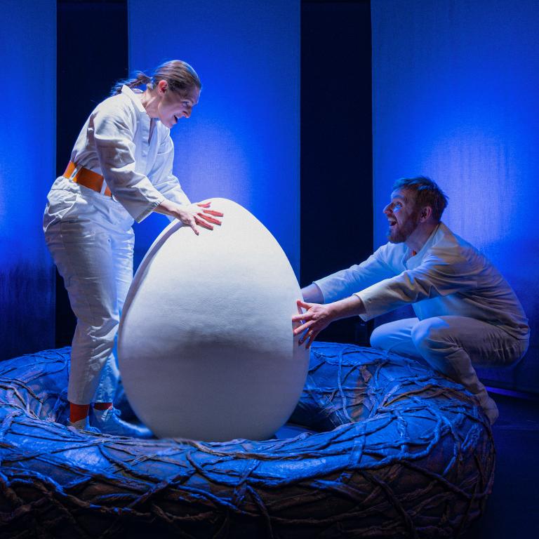 Två personer med vita kläder står i blått ljus och tittar på ett stort ägg