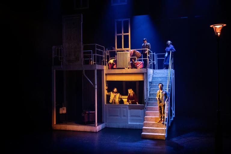 Översiktsbild av scenografi med trappa, balkong och flera personer i olika våningar av ett hus.