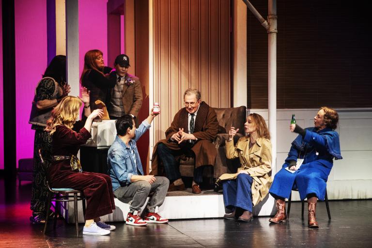 Flera skådespelare sitter och samtalar i en grupp runt en äldre man i en fåtölj på scenen.