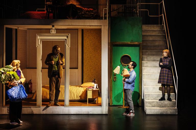 Flera skådespelare står runt scenbilden av en lägenhet med trappa och rum i genomskärning. En håller på en krukväxt en annan i en lampa.