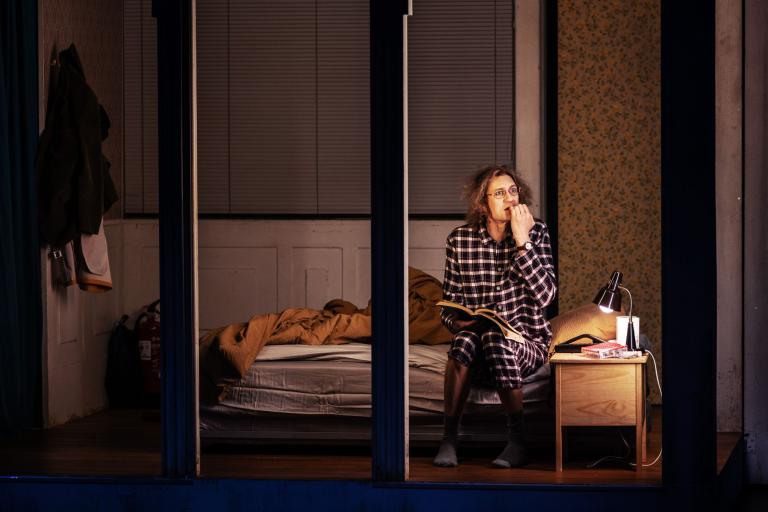 Person sitter på en säng i ett rum och läser en bok vid en liten sänglampa, tuggar nervöst på naglarna..