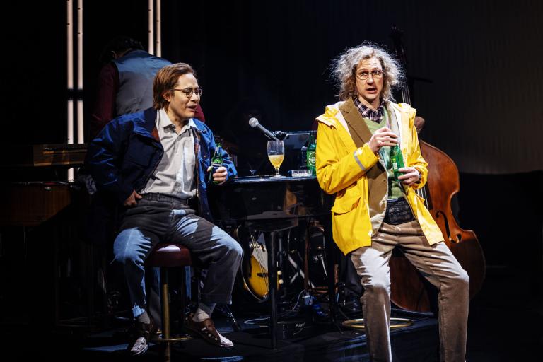 Två personer sitter vid ett bord med mikrofoner och dryck medan en kontrabas syns i bakgrunden.