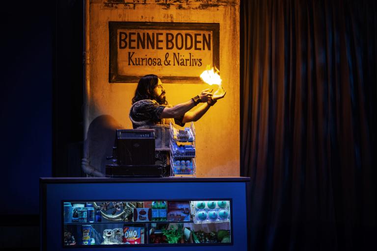 Person står bakom en butiksdisk och trollar fram eld i händerna under skylten “Benneboden Kuriosa & Närliv”.