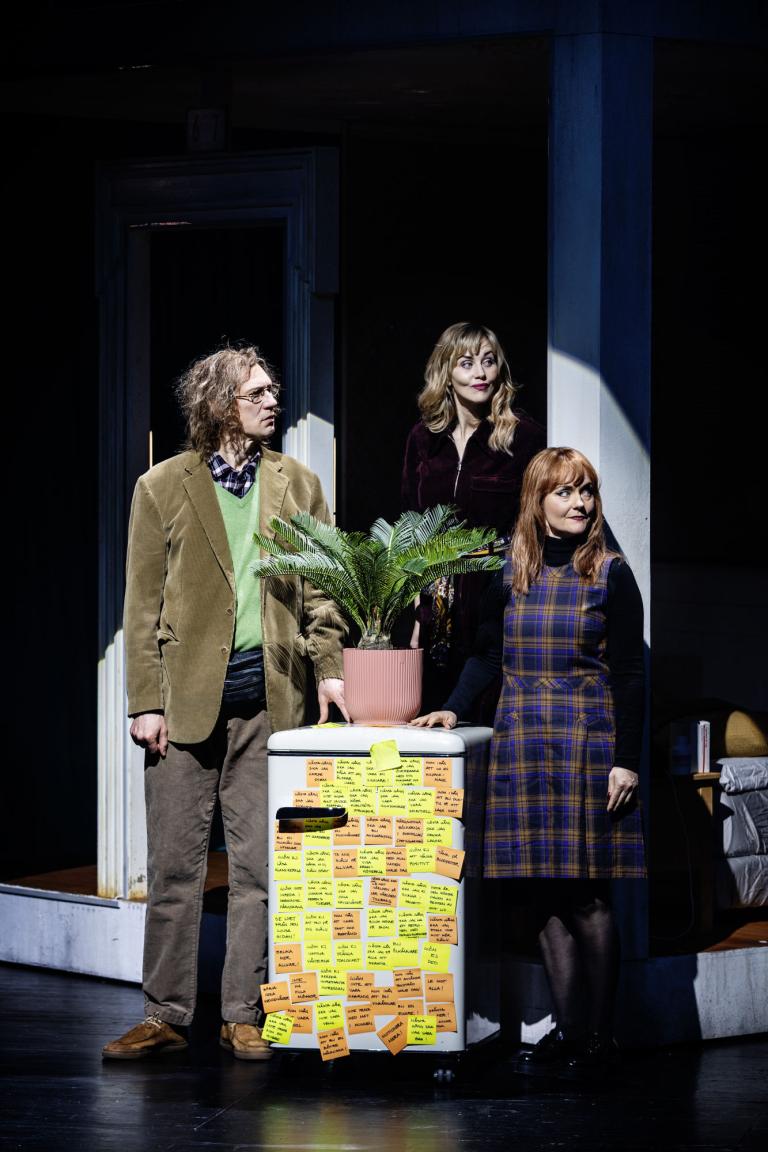 Tre personer står vid ett kylskåp täckt av post-it-lappar med en krukväxt ovanpå.