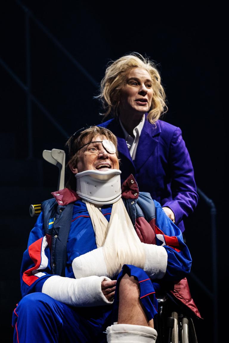Person i rullstol med bandage och nackkrage, bakom står en kvinna i lila kavaj.