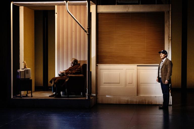 En äldre man sitter i en fåtölj medan en annan person står utanför rummet.