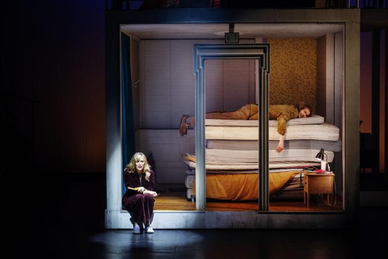 Person ligger på en säng i ett rum medan en annan sitter utanför och skriver i ett block.