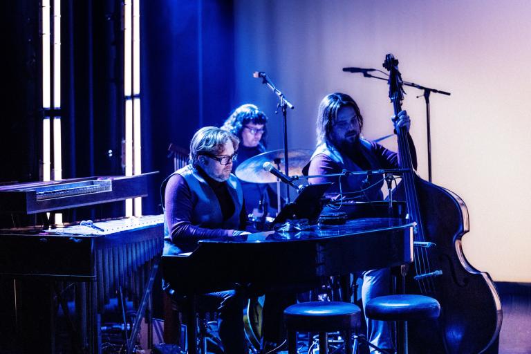Musiker spelar piano och kontrabas och trummor på scenen i blått scenljus.