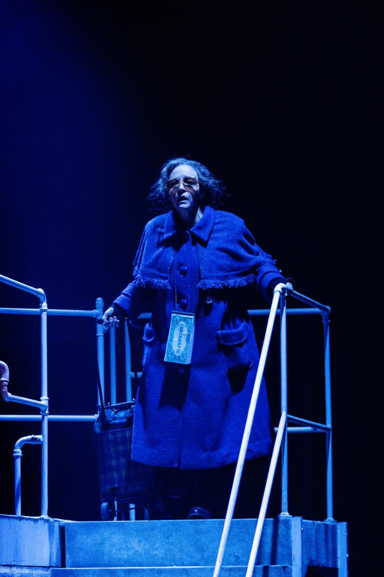 Kvinna i blå kappa står högst upp på en trappa i blått scenljus.