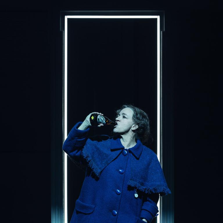 Person i blå kappa dricker ur en flaska framför en lysande dörrkarm på scenen.