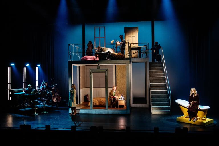 Översiktsbild av scenografi som föreställer ett flervåningshus med rum, trappa och flera skådespelare på scenen. Mörkt blått ljus