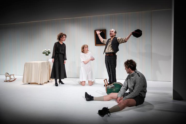 Fyra skådespelare på vit scen; en man står upp och gestikulerar medan en annan sitter på golvet, två kvinnor står och knäböjer bredvid.