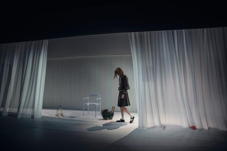 Ensam kvinna går över scenen mellan vita gardiner; barnvagn och stol i bakgrunden.