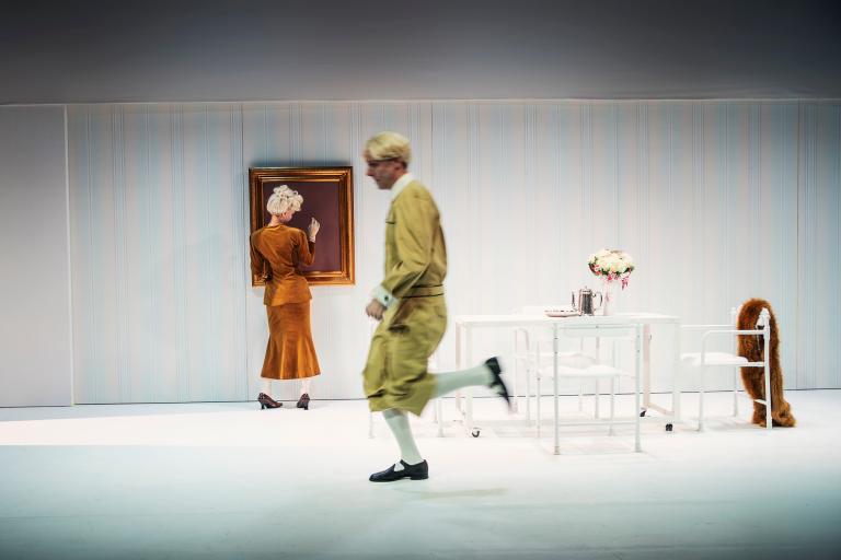 Kvinna betraktar en tom guldram på väggen medan en man passerar förbi; vitt möblemang i rummet.