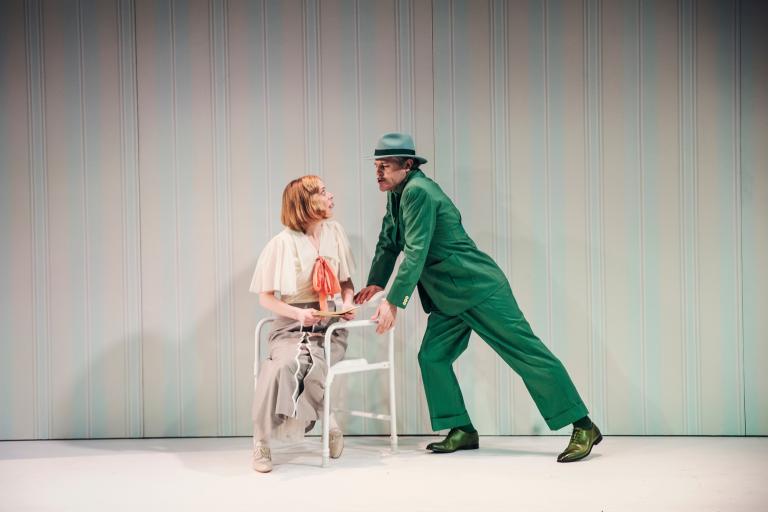 Man i grön kostym lutar sig mot sittande kvinna; de ser intensivt på varandra.