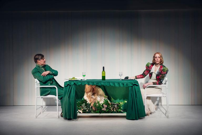 Två personer sitter vid bord med grön duk; en tredje person syns under bordet bland växter.