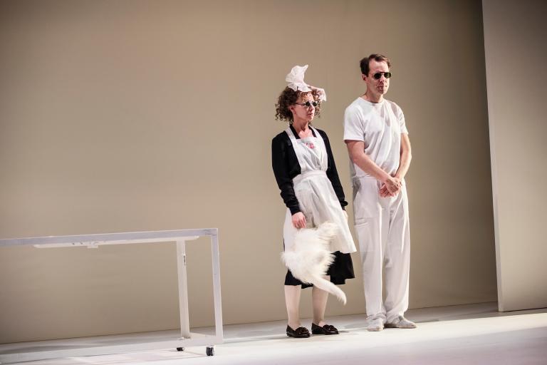 Två personer står bredvid varandra mot neutral bakgrund; en i förkläde håller en vit pälskatt.