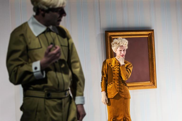 Kvinna i gul dräkt poserar vid tom guldram; en man i förgrunden är ur fokus.