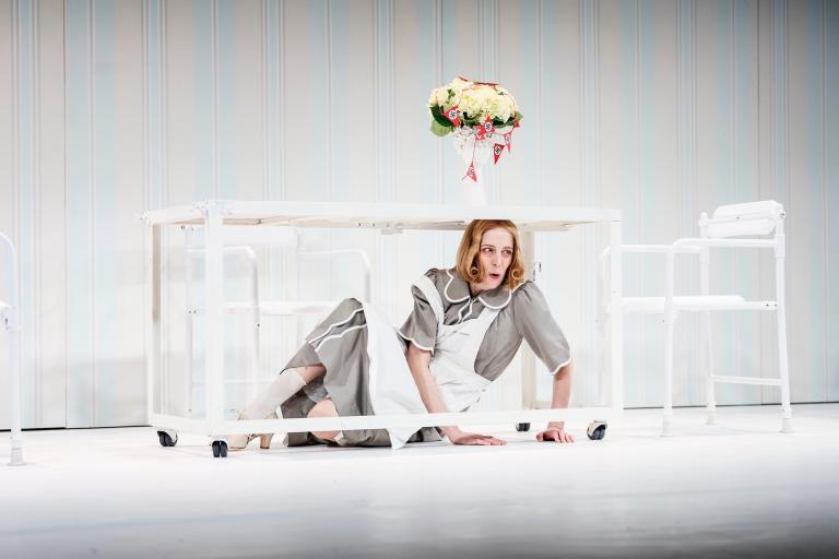 Kvinna kryper under ett bord; en vas med blommor står ovanpå.