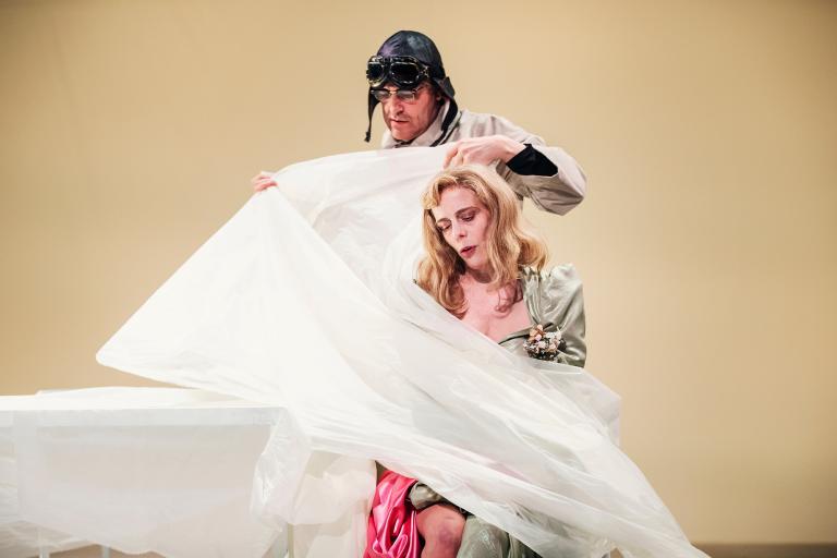 Man i pilotmössa håller upp ett vitt tyg bakom sittande kvinna.