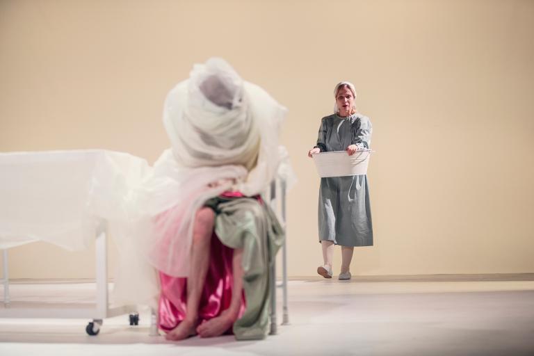 Sjuksköterska går mot en säng där en kvinna sitter insvept i tyg.