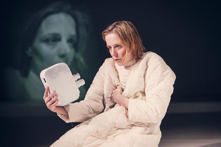 Kvinna i vadderad dräkt håller upp en filmkamera mot sitt ansikte. Hennes ansikte projuceras på väggen bakom..