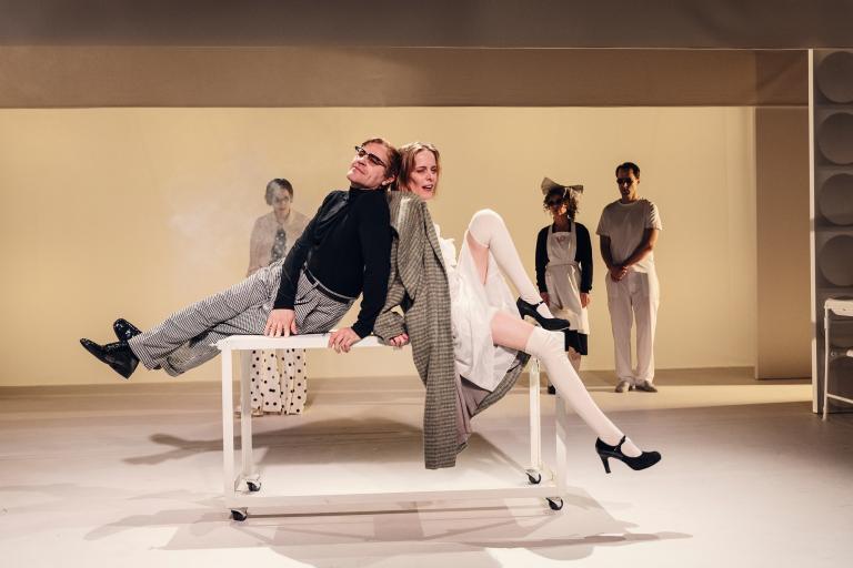 Två skådespelare sitter rygg mot rygg på ett bord; övriga ensemblemedlemmar står i bakgrunden.