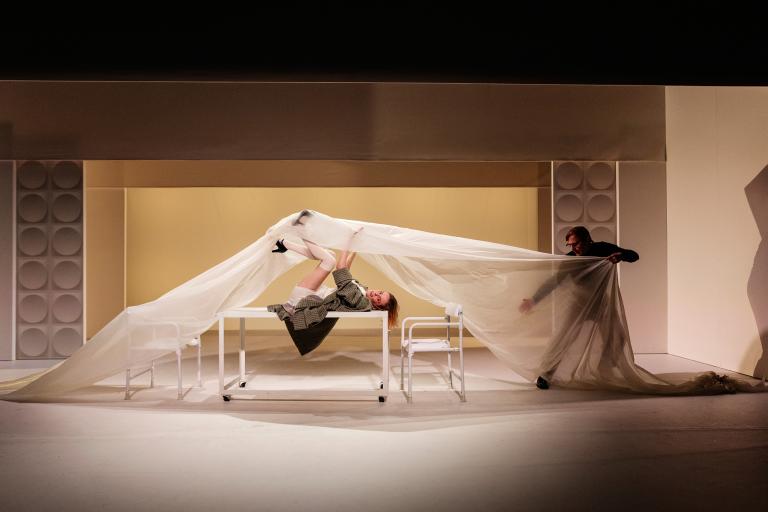 Kvinna ligger på rygg på ett bord medan en man drar ett stort vitt tyg över scenen.