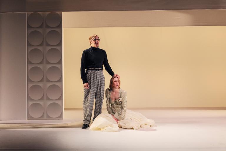 Man står bakom knäböjande kvinna och håller handen på hennes huvud; hon ser upp.