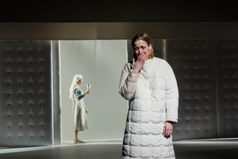 Kvinna i vadderad vit dräkt står i förgrunden och håller handen för munnen; en annan kvinna syns i bakgrunden med en glasburk.