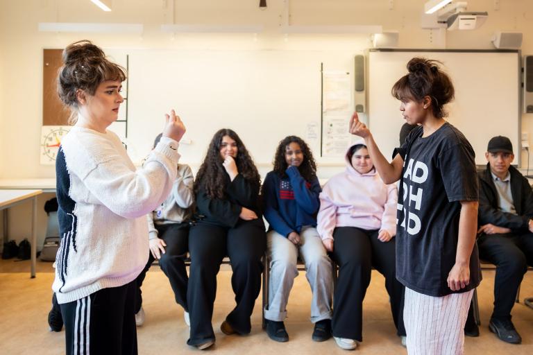 Två skådespelare står mittemot varandra och låtsas som att hålla något litet mellan fingrarna. Elever i bakgrunden