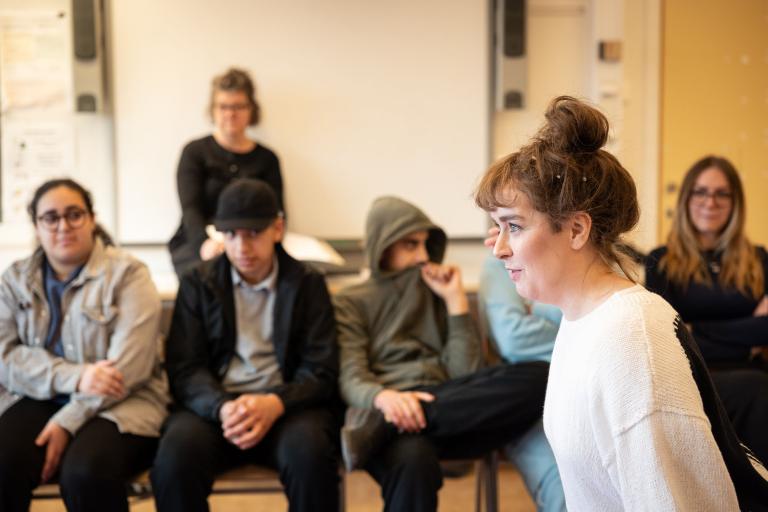 En skådespelare med vit tröja står och tittar intensivt rakt fram. Elever sitter i bakgrunden