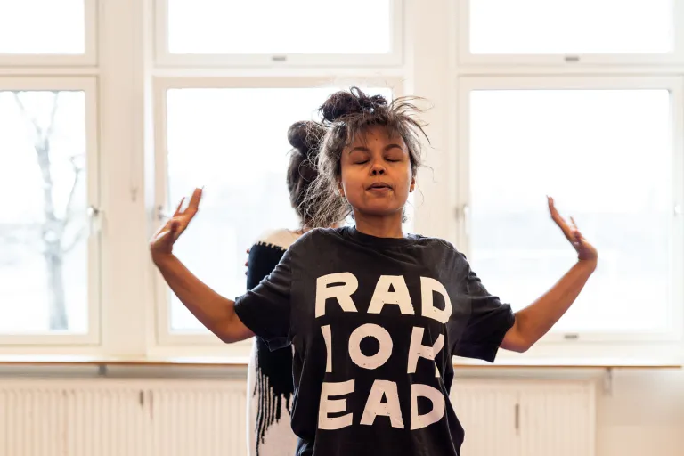 en skådespelare med radiohead-tröja blundar och lyfter händerna utåt. En annan skymtar bakom. Fönster i bakgrunden