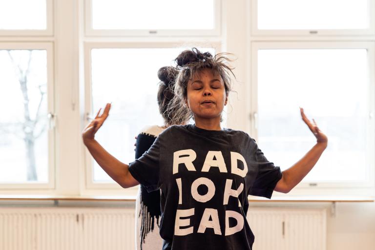 en skådespelare med radiohead-tröja blundar och lyfter händerna utåt. En annan skymtar bakom. Fönster i bakgrunden