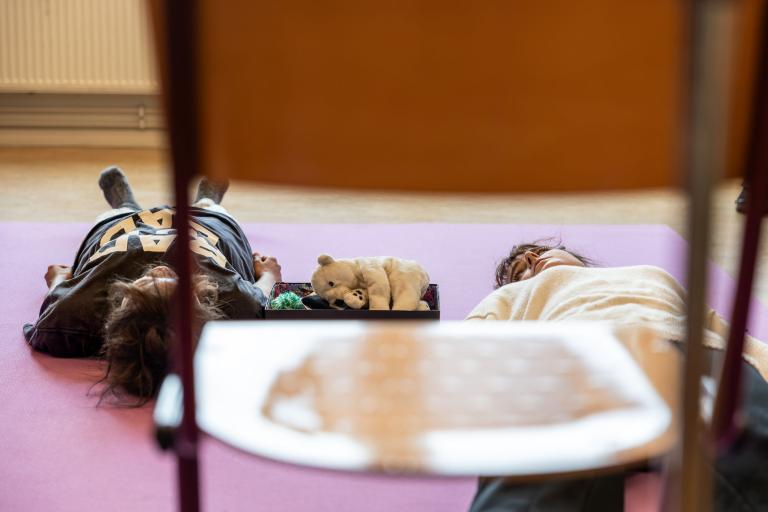 i förgrunden en klassrumsstol. två skådespelare ligger bakom på en rosa matta