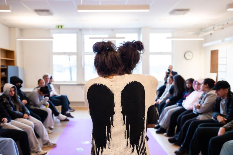 Skådespelare står i ett klassrum med ryggen mot kameran. Stickad tröja med vingar. elever sitter på två rader om en rosa matta