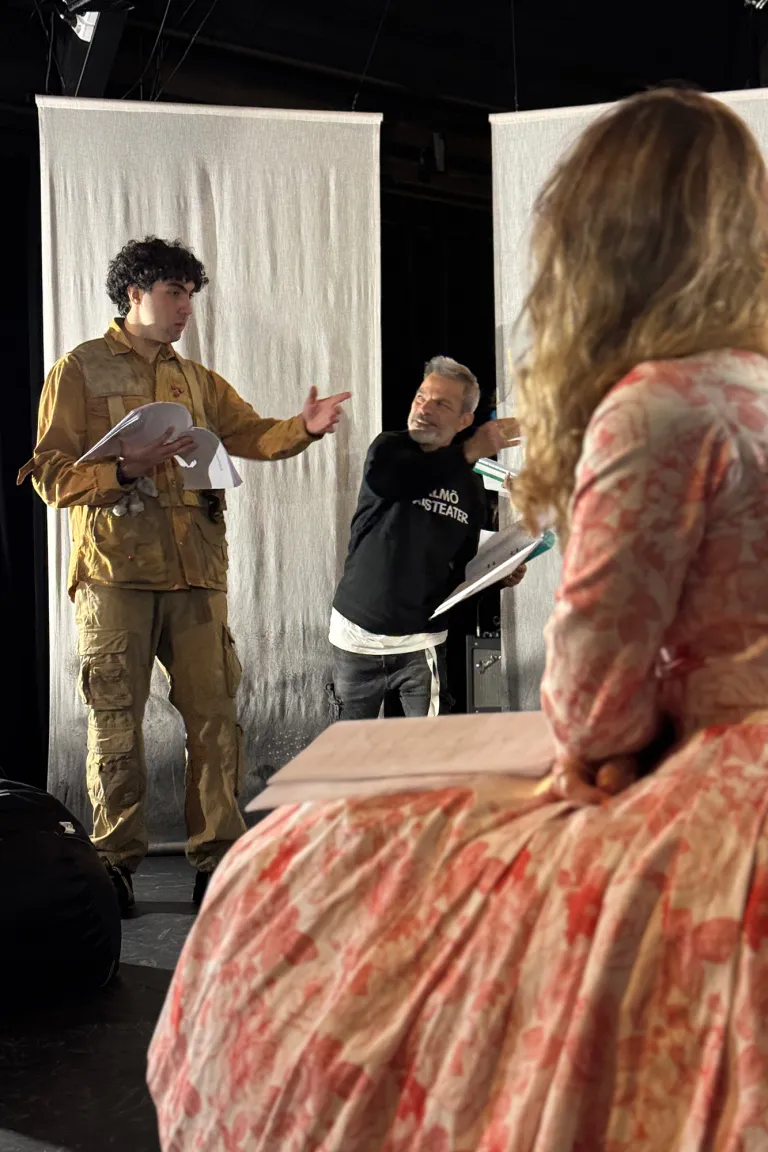En person med svart Malmö Stadsteater-tröja står och pratar med en ung person med militärkostym. I förgrunden sysn en annan person med blommönstrad klänning