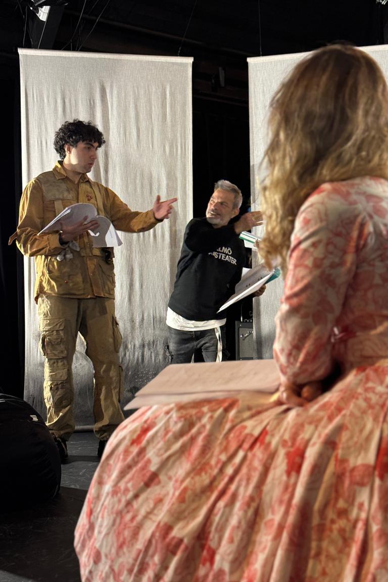 En person med svart Malmö Stadsteater-tröja står och pratar med en ung person med militärkostym. I förgrunden sysn en annan person med blommönstrad klänning