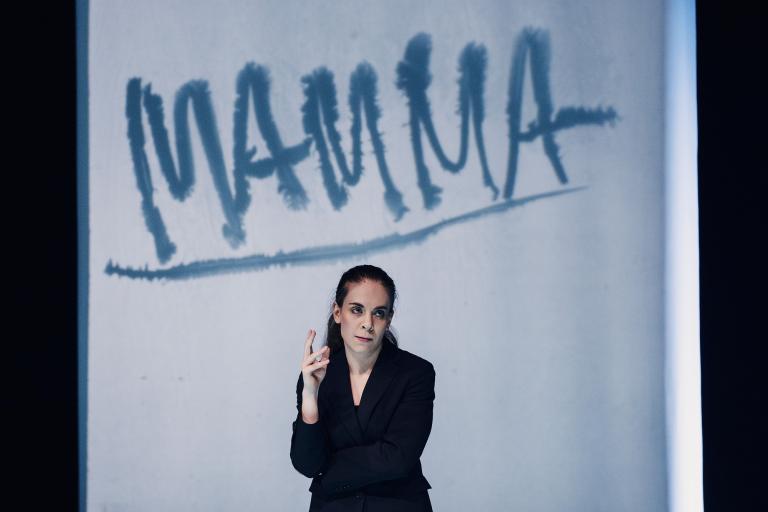 Skådespelare i kostym framför projektion med texten ”Mamma”.