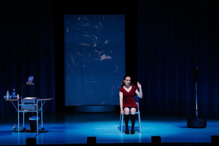 Skådespelare sitter på stol på tom scen med projektion bakom.