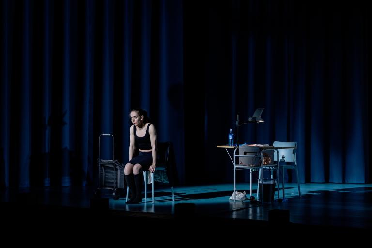 Skådespelare sitter framåtlutad på stol i mörk scenbild.