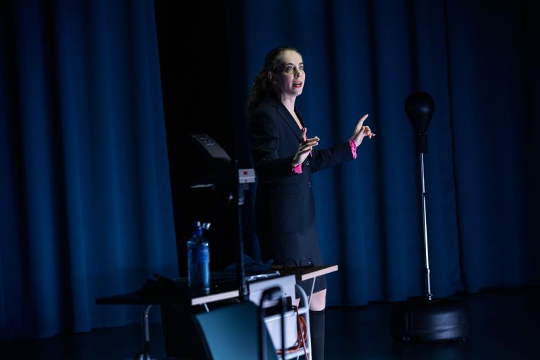 Skådespelare talar stående vid bord med teknik på scen.