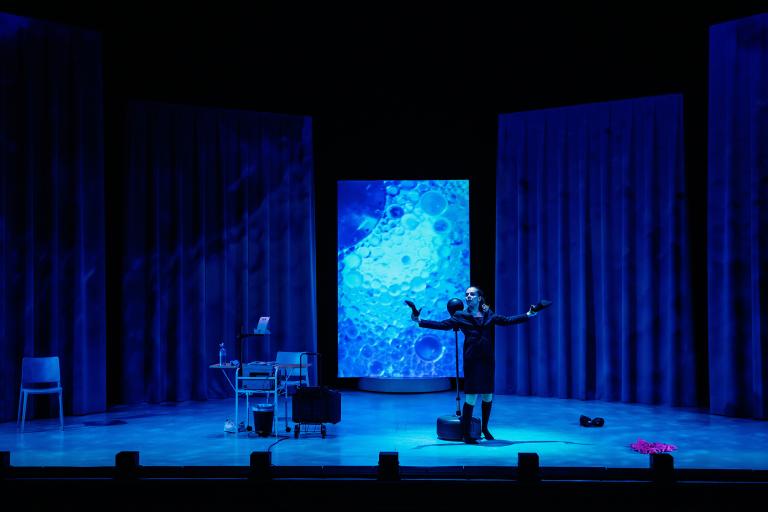Skådespelare i helbild på scen med blå projektioner.