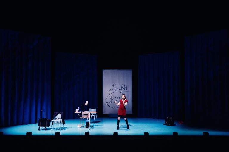 Skådespelare står centralt på scen med textprojektion bakom.