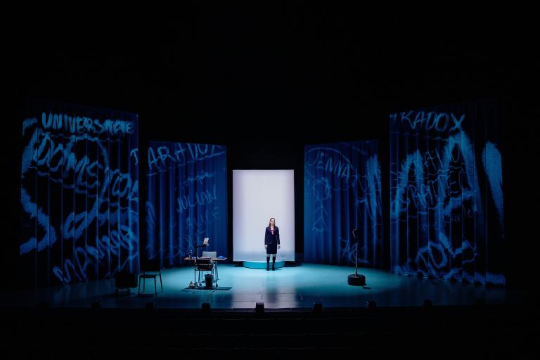 Skådespelare står ensam i upplyst scenrum med projektioner.